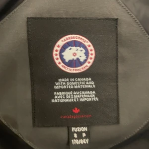 Grå Canada Goose dunjacka med päls - Säljer en grå dunjacka från Canada Goose med klassisk patch på ärmen och stor huva med päls. Jackan har dragkedja, knappar och ribbade muddar. Perfekt för kalla dagar och riktigt snygg streetstil.          Frågor? Tveka inte på att skriva!