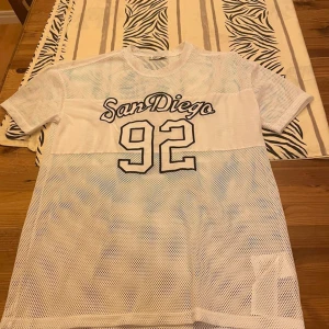 Vit mesh t-shirt San Diego FB Sister - Vit mesh t-shirt från FB Sister med trycket 'San Diego 92' på bröstet. Oversized känsla, kortärmad och tillverkad i luftig polyester. Perfekt för en sportig och trendig look.