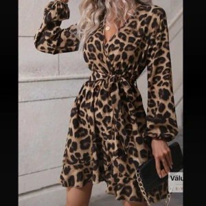Leopardmönstrad omlottklänning - Snygg kort leopardmönstrad omlottklänning med långa ärmar och knyt i midjan. Klänningen har volangdetaljer nertill och v-ringad hals. Perfekt för dig som vill sticka ut med ett djurmönster och en feminin siluett.