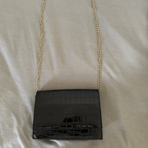 Svart axelväska med kedja - Snygg svart axelväska med krokopräglat mönster och guldfärgad kedja som axelrem. Väskan har ett stilrent och elegant utseende med magnetlås och flera fack inuti. Perfekt för dig som vill ha en trendig och praktisk väska till mobilen och småsaker.