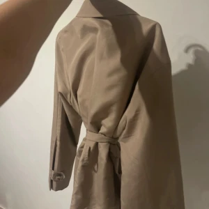 Beige trenchcoat från Vero Moda - Snygg beige trenchcoat från Vero Moda med klassisk krage, knappar framtill och bälte i midjan. Jackan har långa ärmar och en stilren look som passar perfekt till vår och höst. Materialet är mjukt och lätt, vilket gör den enkel att bära.