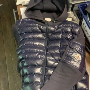 Mörkblå pufferjacka från Moncler - Snygg mörkblå pufferjacka från Moncler med glansig quiltad front, huva och ärmficka med logotyp. Jackan har dragkedja framtill och mjuka ärmar och rygg i bomullsmix. Perfekt för dig som vill ha en sportig och exklusiv look.