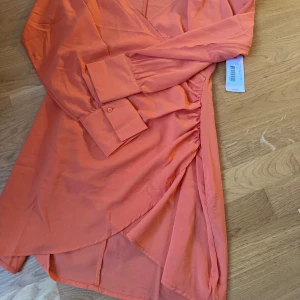 Orange omlottklänning från Nelly - Snygg orange kortklänning från Nelly med omlott-design och långa ärmar. Klänningen har knappar vid ärmsluten och en cool rynkad detalj i sidan. Perfekt för dig som gillar färg. 