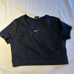 Svart croppad träningstopp från Nike - Säljer en svart croppad träningstopp från Nike med korta ärmar och rund halsringning. Toppen har en liten vit Nike-logga framtill och ribbade detaljer på ryggen för extra stil. Perfekt för gymmet eller sportiga outfits.