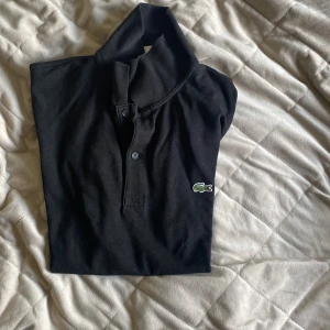 Svart pikétröja från Lacoste - Svart pikétröja från Lacoste med klassisk krage, två knappar och den ikoniska gröna krokodilloggan på bröstet. Tröjan är i mjuk bomull och har korta ärmar. Perfekt för en clean och sportig look.