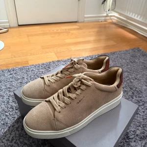 Beige mockasneakers från Gant strl 45 - Snygga sneakers från Gant i beige mocka med vita sulor och orange detaljer på snörningen. Skorna har rund tå, platt sula och cognacsfärgad detalj vid hälen. Perfekta för dig som gillar stilrena och klassiska sneakers med en modern touch.