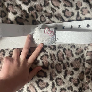 Hello Kitty bälte  - Superfint vitt bälte från Hello Kitty med bred rem i syntet och stor silvrig Hello Kitty-spänne med rosa detaljer. Perfekt accessoar för dig som älskar Sanrio och vill sticka ut med något unikt och gulligt. Har en svart fläck som visas på bilden. 98cm. 