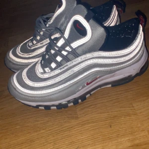 Nike Air Max 97 Silver Bullet - Nike Air Max 97 Silver Bullet med ikoniska vågformade linjer i silver och grått, röda swoosh-detaljer och svart sula. Skorna har reflekterande detaljer, snörning och synlig Air Max-dämpning. Perfekta för dig som vill sticka ut med en klassisk sneaker.