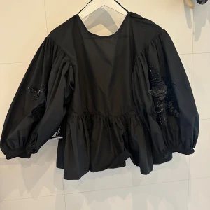 Svart blus med volymärmar och broderi från HM - Säljer en svart blus med voluminösa ärmar och broderade blommor i svart paljett på båda ärmarna. Blusen har rund halsringning, rynkad midja och elastiska ärmslut. Perfekt för dig som gillar unika detaljer och statement-ärmar.