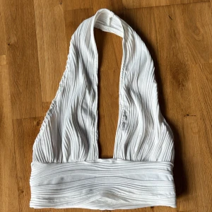 Vit ribbad halterneck topp - Säljer en vit ribbad halterneck topp med djup urringning och bred resår nedtill. Toppen är ärmlös och har en snygg struktur i tyget som ger en cool look. Perfekt att styla med jeans eller kjol för en trendig outfit.