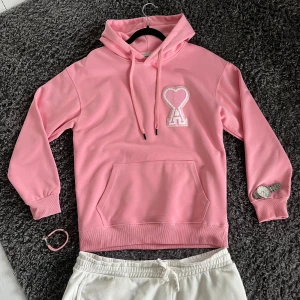 Ami alexandre mattiussi - Säljer en rosa hoodie från Ami Alexandre Mattiussi med broderad hjärtlogga och bokstaven A på bröstet. Tröjan har huva med snörning, magficka och långa ärmar. Perfekt för dig som vill ha en snygg och bekväm streetstyle-look.