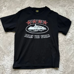 Svart kortärmad t-shirt Corteiz S - Svart t-shirt från Corteiz med tryck på bröstet där det står 'Corteiz Rules The World' och en stor grafisk illustration. Klassisk passform och rund hals. Tillverkad i 100% bomull för en skön känsla. Använd 1 gång! 