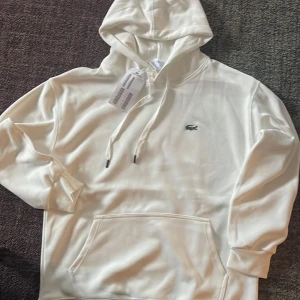 Vit hoodie från Lacoste med huva - Snygg vit hoodie från Lacoste med klassisk krokodillogga på bröstet. Tröjan har huva med snörning, känguruficka och ribbade muddar. Regular fit och tillverkad i mjuk bomull för en skön känsla. Perfekt för dig som gillar stilrena och sportiga plagg.