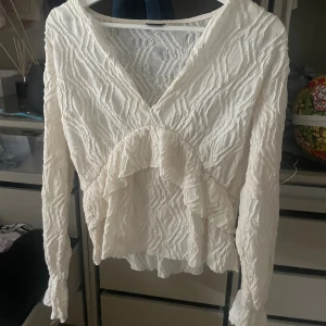 Vit volangblus från Gina Tricot - Supersöt vit blus från Gina Tricot med crinklad struktur och volangdetalj framtill. Blusen har v-ringning och långa ärmar, perfekt för dig som gillar romantisk och trendig stil. Materialet är lätt och luftigt, vilket gör den skön att bära.💞💞💞