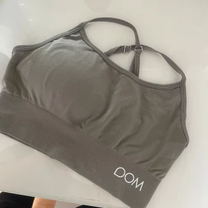 Grå sporttopp från Drop of Mindfulness - Säljer en grå sporttopp från Drop of Mindfulness med smala justerbara axelband som korsas i ryggen. Toppen har bred ribbad kant nedtill och logga både fram och bak. Perfekt för träning och ger en snygg, sportig look.