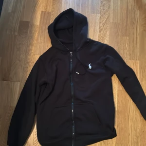 Svart hoodie från Ralph Lauren - Svart hoodie från Ralph Lauren med dragkedja och huva. Klassisk vit logga på bröstet och snörning i huvan. Mjuk och skön, perfekt för chill dagar. Stora fickor framtill och stilren look.