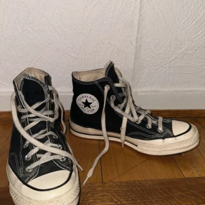 Converse Chuck Taylor All Star svart - Klassiska Converse Chuck Taylor All Star i svart canvas med vita detaljer och snörning. Högt skaft, rund tå och vit gummisula med svart rand. Ikonisk logga på sidan och tydliga kontrastsömmar. Perfekta för en cool och avslappnad stil.