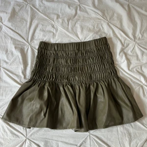 Olivgrön kort kjol från Continue - Supersnygg olivgrön kort kjol från Continue i skinnimitation. Kjolen har smockad midja som ger en cool och voluminös effekt, samt en lätt utsvängd form. Perfekt för dig som gillar trendiga detaljer och vill sticka ut med en unik look.