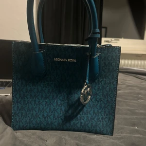 Blå handväska från Michael Kors - Snygg blå handväska från Michael Kors med MK-logga i silver och mönstrad yta. Väskan har två handtag, dragkedja upptill och flera fack inuti. Tillverkad i skinn med detaljer i matchande blått. Perfekt för dig som vill ha en stilren och trendig accessoar.