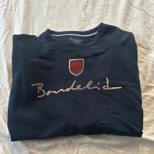 Mörkblå sweatshirt från Bondelid - Mörkblå sweatshirt från Bondelid med broderad logga och röd-svart sköld på bröstet. Klassisk rund halsringning och mjukt material som känns skönt mot huden. Perfekt för en avslappnad och stilren look.