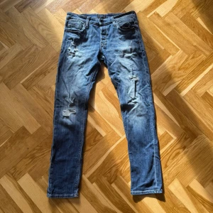 Jack and Jones blå slitna jeans - Säljer ett par blå slim jeans från Jack and Jones med slitningar och hål på benen. Storlek W32/L34