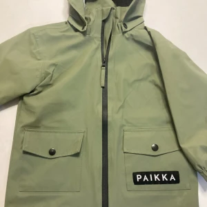 Grön regnjacka från PAIKKA med huva - Snygg grön regnjacka från PAIKKA med huva och stora fickor framtill. Jackan har svart dragkedja och tryckknappar på fickorna. Insidan är fodrad med grått tyg med hundmönster. Perfekt för regniga dagar och streetstyle.