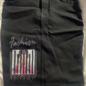 Svart kortkjol med strass och tryck - Svart kortkjol i jeansstil med två bakfickor och bälteshällor. Framsidan har ett coolt tryck med motiv och texten 'Fashion' i strass samt dekorativa stenar. Kjolen har rak passform och dragkedja framtill.