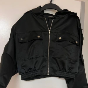 Svart croppad bomberjacka PrettyLittleThing - Svart croppad bomberjacka från PrettyLittleThing med huva, stora fickor framtill och silvrig dragkedja. Jackan har elastisk midja, långa ärmar med detaljerade knappar och ringar i metall. Snygg oversized passform och glansig finish.