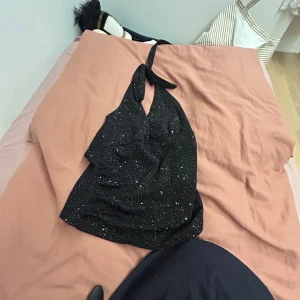 Svart glittrig halterneck topp - Supersnygg svart halterneck topp med glittrigt tyg och knyt i nacken. Toppen har en croppad passform och är perfekt för dig som vill sticka ut. Materialet är mjukt och stretchigt, vilket gör den bekväm att bära hela kvällen.Aldrig använd funkat oxå både S och M. Den är fån Temu 