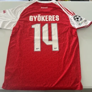 Arsenal Gyökeres 14 matchtröja Adidas - Snygg Arsenal fotbollströja från Adidas med Gyökeres och nummer 14 på ryggen. Röd bas med vita ärmar, broderat klubbmärke, Champions League-patch och klassiska Adidas-ränder. Tillverkad i lätt och ventilerande polyester med subtilt mönster över hela framsidan. Replika, endast handtvätt gäller!