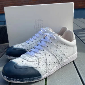 Maison Margiela sneakers vit/svart - Maison Margiela sneakers i vitt skinn med svart färgstänk och mörkblå detaljer på tån. Klassisk låg modell med vit platt sula och vita snören. Unik design med handmålat stänkmönster som ger en cool och modern vibe.