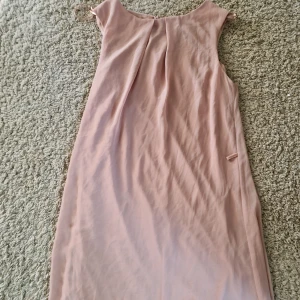 Ljusrosa klänning från Atmosphere - En stilren ljusrosa klänning från Atmosphere i rak modell. Klänningen är ärmlös och har en enkel design med diskreta veck framtill och knytband vid axlarna. Perfekt för dig som gillar minimalistisk och feminin stil. Passar S/M