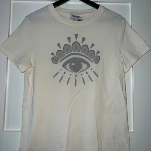 Vit Kenzo t-shirt - Snygg vit t-shirt från Kenzo i bomull med ikoniskt ögontryck framtill. Klassisk rund halsringning och korta ärmar. Perfekt för dig som vill sticka ut med en stilren men unik look. I mycket gott skick och knappt använd! 