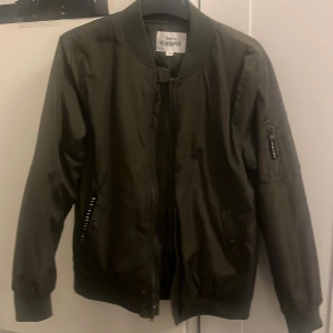 Olivgrön bomberjacka från KappAhl - Snygg olivgrön bomberjacka från KappAhl med klassisk ribbad krage och muddar. Jackan har dragkedja framtill, fickor med dragkedja och en extra ficka med zip på ärmen. Tillverkad i glansig polyester som ger en cool look.