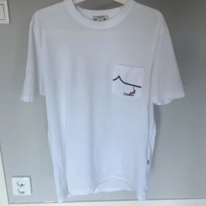 Vit t-shirt från Jacob Cohën - Vit t-shirt från Jacob Cohën med en liten bröstficka som har ett stilrent tryck av en linje och en röd detalj. Klassisk rund hals och avslappnad passform. Tillverkad i mjuk bomull, perfekt för en clean och enkel look. Köpt på united fashion i Göteborg för 2900kr helt ny med lappar.