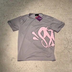 Syna world  - Snygg grå t-shirt från Syna World  med stort rosa grafiskt tryck på framsidan. Klassisk rund hals och korta ärmar. Perfekt för dig som gillar streetstyle och vill sticka ut med en cool detalj på bröstet.