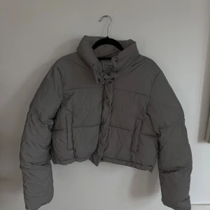 Reflex pufferjacka NA-KD - Grå croppad pufferjacka från Hannalicious x NA-KD med hög krage och dragkedja framtill. Jackan är i reflex. Jackan har två fickor på sidorna och quiltad design. Perfekt för kalla dagar när du vill ha en stilren och trendig look.