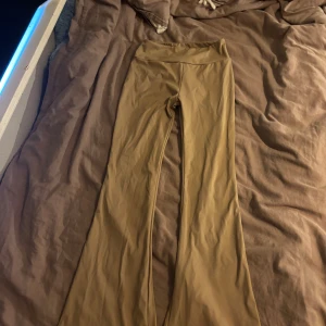 Beiga bootcut byxor med fickor - Snygga beiga bootcut byxor med hög midja och två bakfickor med knappar. Byxorna har en lätt glansig finish och är gjorda i ett stretchigt syntetmaterial. Rynkdetalj bak ger extra form. Perfekta för dig som gillar trendiga och bekväma byxor.