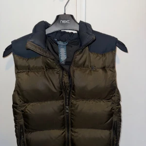 Olivgrön dunväst från Polo Ralph Lauren - Snygg olivgrön dunväst från Polo Ralph Lauren med svarta axelpartier och dragkedja framtill. Västen har hög krage och två fickor med dragkedja. Broderad logga på bröstet. Är i väldigt bra skicke och väldigt skön att ha. Är lite tjockare än normala västar men inte speciellt mycket. Priset är ej hugget i sten. Är barn size och passar ca 154cm.