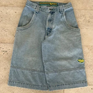Baggy jeansshorts från Arlo - Baggy ljusblå jeansshorts från Arlo med grönt och gult broderat märke på benet och bakfickan. Shorts med bred passform, stora fickor och råa kanter nertill. Klassisk femficksdesign och bälteshällor. Perfekta för en avslappnad streetstil.