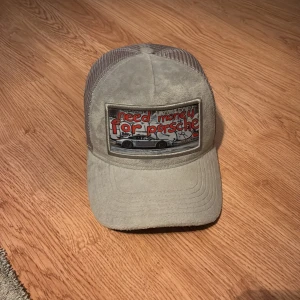 Grå keps från Revenite med Porsche-tryck - Säljer en grå keps från Revenite med mesh på sidorna och baksidan. Framtill finns ett stort tygmärke med texten 'need money for porsche' och en bild på en Porsche. Kepsen har böjd skärm och justerbar passform. Perfekt för dig som gillar bilar och streetstyle.