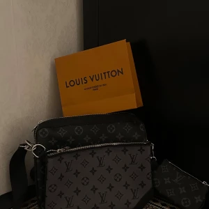 Axelväska monogram svart - Snygg axelväska i svart och grått skinn med klassiskt monogrammönster. Väskan har flera fack med dragkedja och justerbar axelrem. Perfekt för dig som vill ha en stilren och lyxig accessoar med ikonisk design.