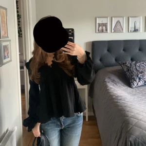 Svart blus med spetsdetaljer - Säljer en svart blus med v-ringning och fina spetsdetaljer vid halsen. Blusen har långa, lite puffiga ärmar med volang vid ärmslut och en lös passform som ger en chill vibe. Perfekt att styla med jeans eller kjol för en snygg look.