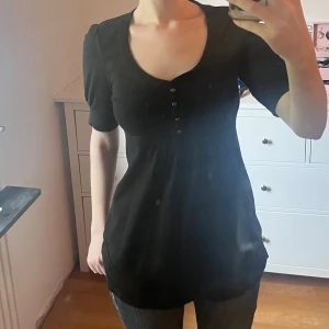 Svart babydoll blus med knappar - Svart blus med rundad urringning och fyra dekorativa knappar framtill. Modellen har halvlånga ärmar och lätt puff vid axlarna. Mjuk och skön bomullskvalitet, lätt utställd nertill för en avslappnad look.