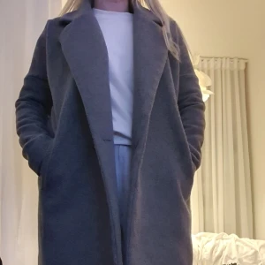 Grå ullkappa med klassisk krage xs - Stilren grå kappa i ullmix med klassisk krage och raka linjer. Kappan har långa ärmar och sidofickor, perfekt för en enkel men snygg look. Passar till det mesta och ger en tidlös vibe. Något snäv i armar, rekommenderar inte att ha tjocktröja med huva under tex