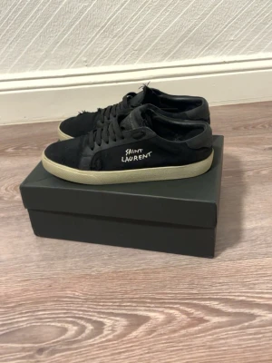  rejäla svarta Saint Laurent sneakers - Svarta sneakers från Saint Laurent med vit sula och handskriven logga på sidan. Skorna har klassisk snörning och är tillverkade i mocka och skinn. Perfekta för dig som gillar en stilren men edgy look.