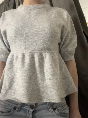 Ljusgrå peplumstickad tröja - Supersöt ljusgrå stickad tröja med peplumskärning och halvlånga ärmar. Tröjan har rund halsringning och ribbade muddar vid ärmslut. Perfekt för dig som gillar en avslappnad sophostyle med en twist.
