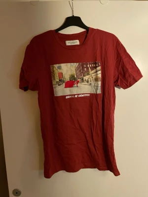 Röd t-shirt från Stock & Hank - Cool röd t-shirt från Stock & Hank med tryck på bröstet som visar en stadsscen och texten 'Santa is coming!'. Klassisk rund hals och korta ärmar. Perfekt för dig som vill sticka ut med en unik print. Tillverkad i mjuk bomull.