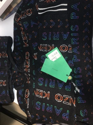 Svart Kenzo hoodie med färgglatt tryck - Cool svart hoodie från Kenzo med färgglada bokstäver som bildar 'KENZO PARIS' över hela plagget. Hoodien har huva med dragsko och ribbade muddar. Perfekt för dig som vill sticka ut med en unik och trendig look.