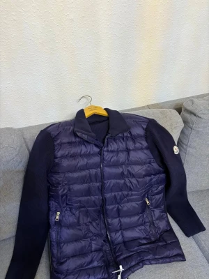 Mörkblå dunjacka från Moncler - Snygg mörkblå dunjacka från Moncler störlek L med quiltad front och stickade ärmar. Jackan har dragkedja framtill, två fickor med dragkedja och klassisk Moncler-logga på ärmen. Perfekt för kyliga dagar och riktigt stilren look.